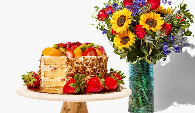 A decadent Strawberry and fruit cake sits on a platter next to a bouquet of fresh flowers
