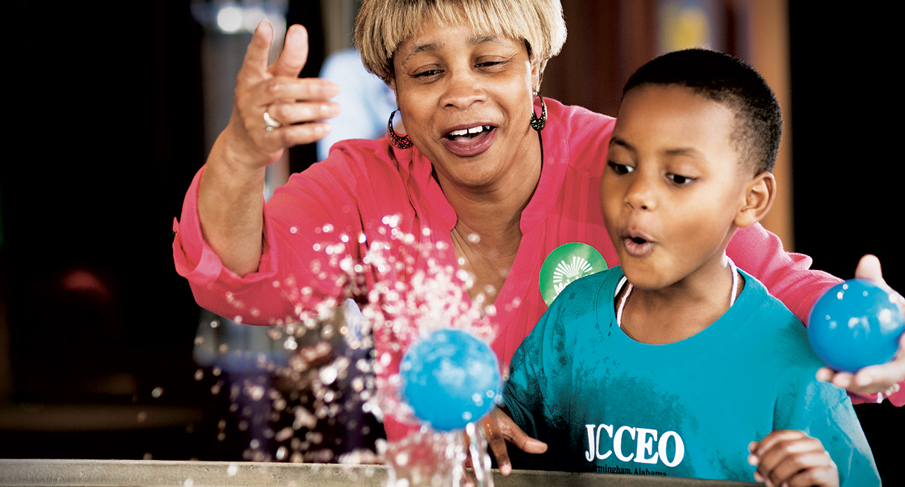 lady teaches educational lesson to youth boy while tossing blue balls into a fountain