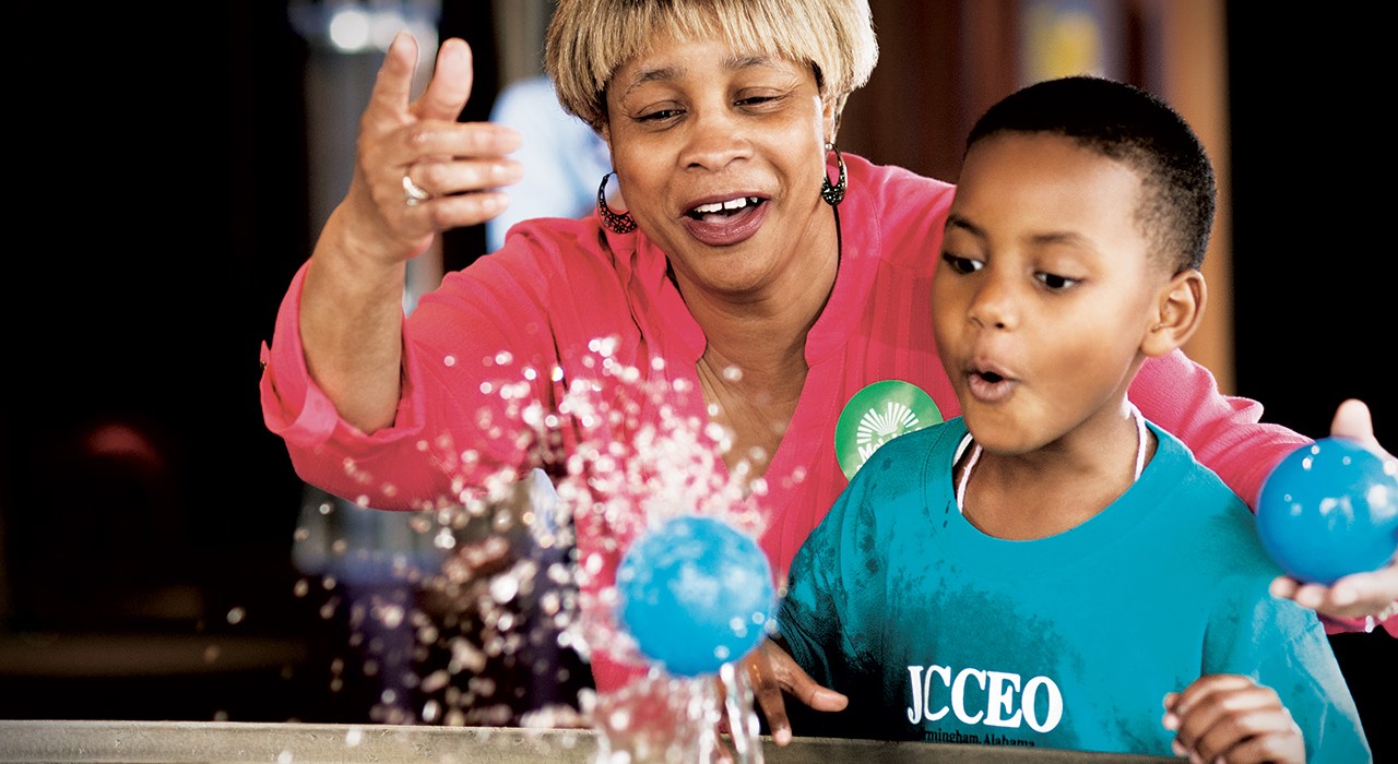 lady teaches educational lesson to youth boy while tossing blue balls into a fountain