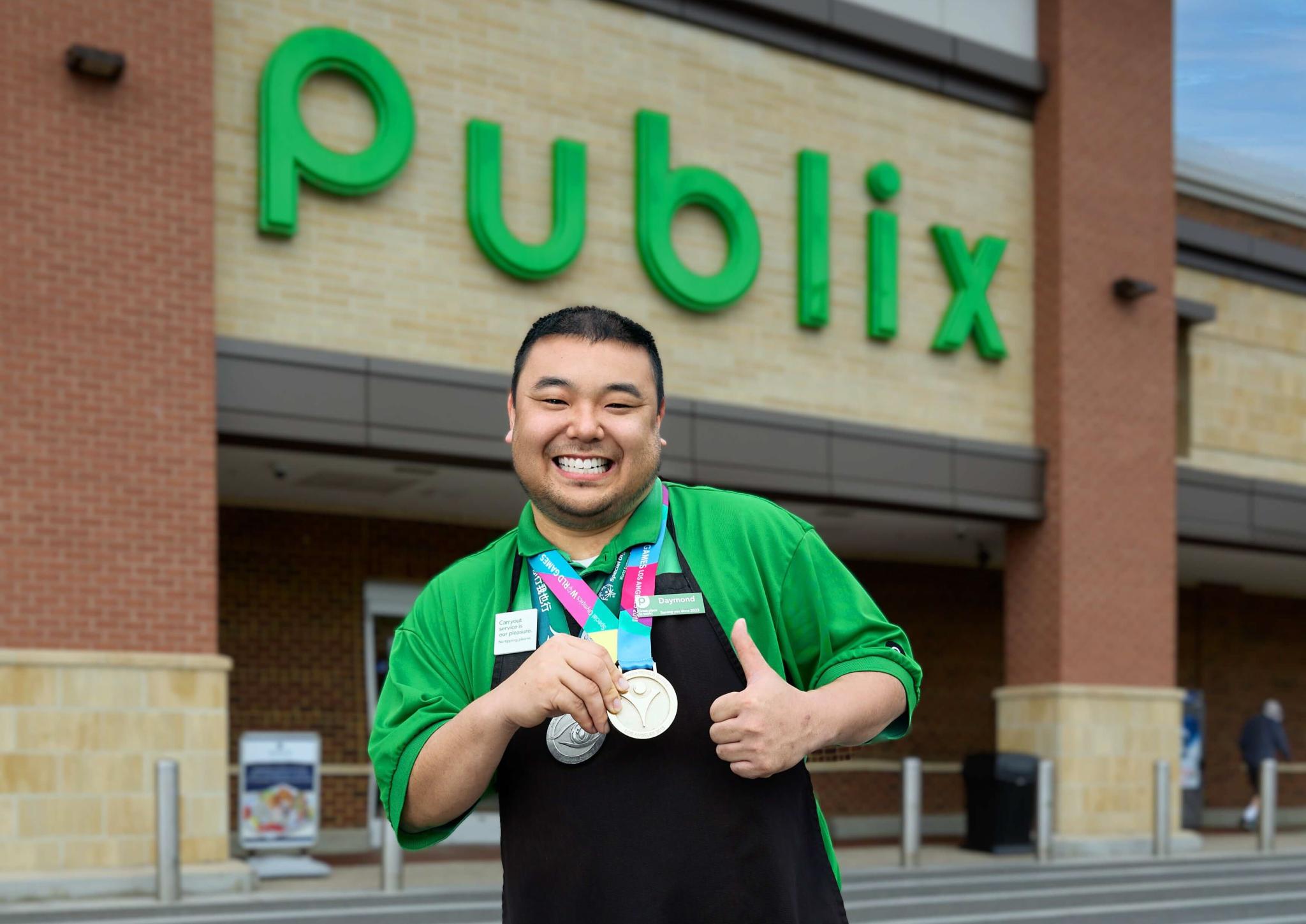 A Publix associate posing outside a Publix storefront with two medals.
