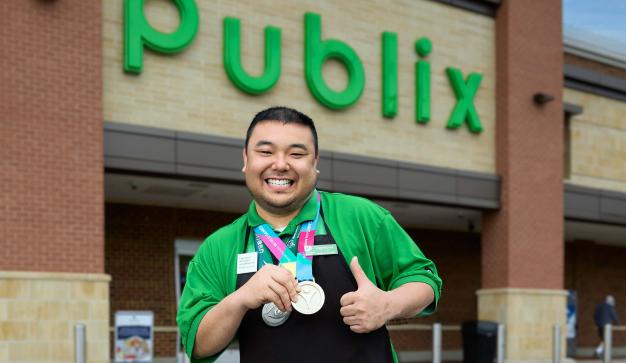 A Publix associate posing outside a Publix storefront with two medals.