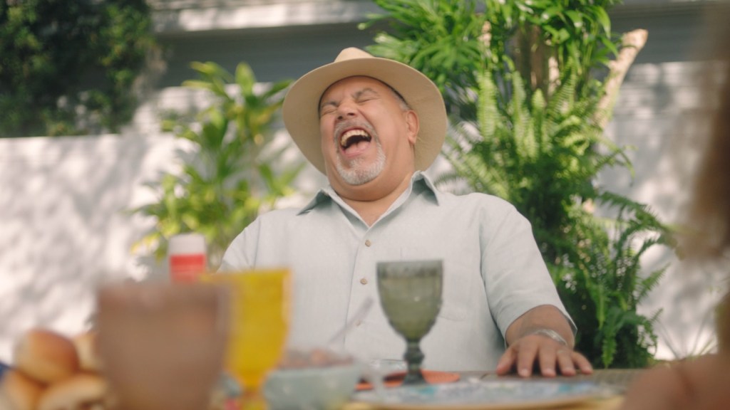Man in hat laughing while sitting at a backyard table filled with food.